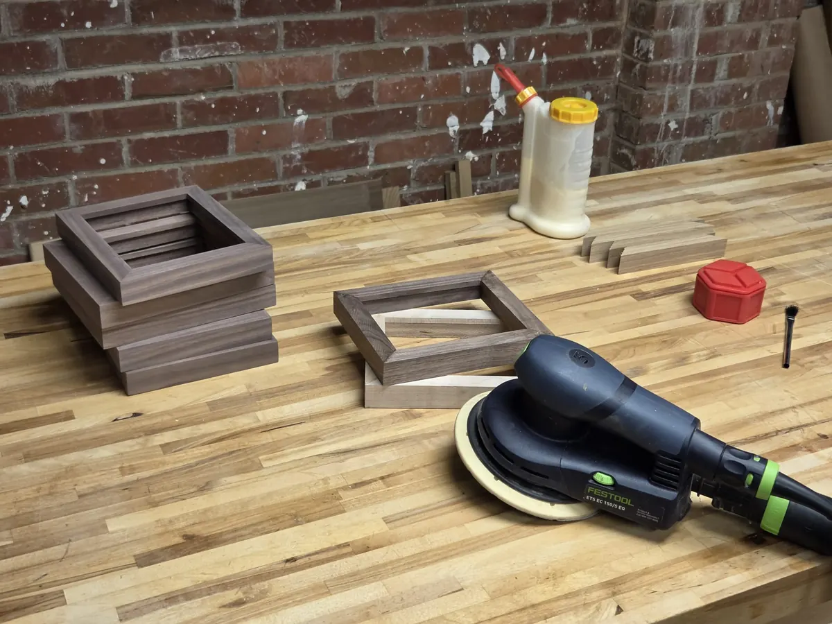 A workshop bench of butcher block against a brick wall: a stack of finished walnut frames, one part-assembled, an orbital sander, glue and a brush